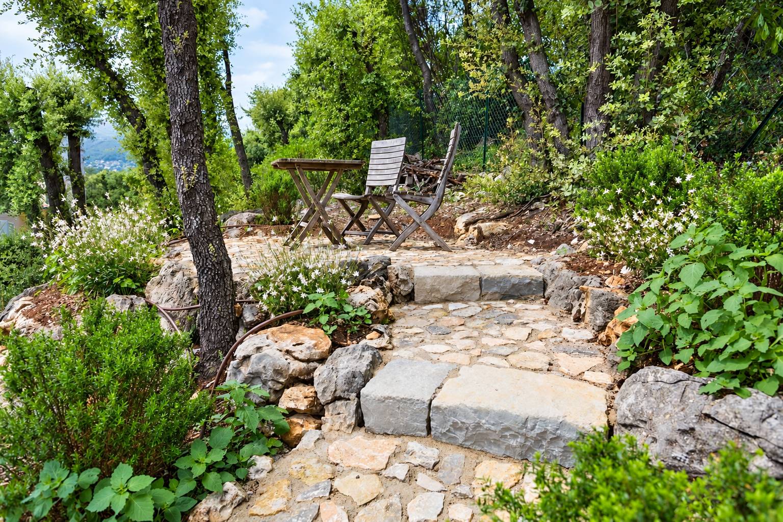 Un escalier en pierres plates mène à une table et deux chaises en bois, au milieu d'un jardin méditerranéen planté d'arbustes et de fleurs blanches.