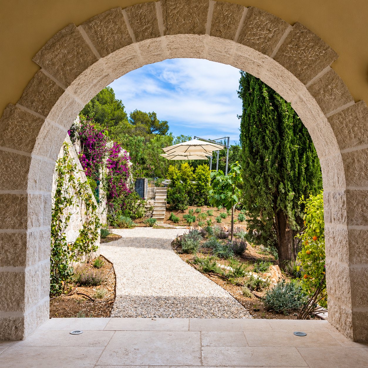 Une arche en pierre claire ouvre sur un jardin méditerranéen avec allée de gravier, cyprès, bougainvilliers et parasol.