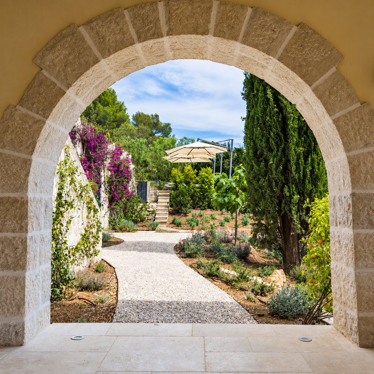 Une arche en pierre claire encadre une allée de gravier bordée de plantes méditerranéennes, de cyprès, de bougainvillées et d'un parasol déporté.