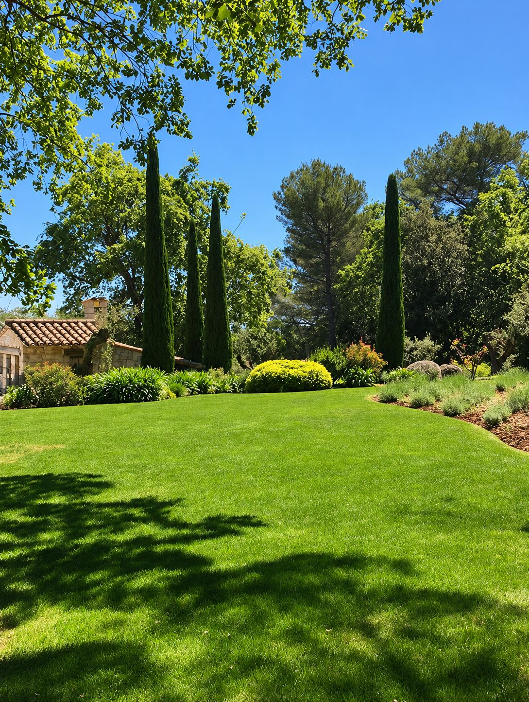 Un jardin ensoleillé avec une grande pelouse verte, des cyprès élancés, des arbustes taillés et une maison en pierre à toit de tuiles.