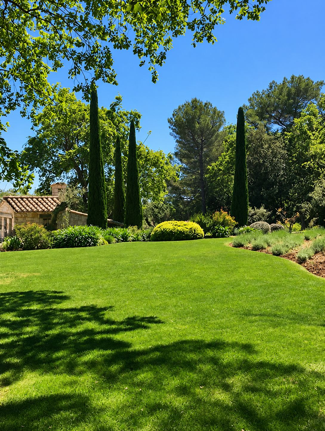 Un grand jardin paysager avec une pelouse verte, des cyprès élancés, des massifs et une maison en pierre à toit de tuiles sous un ciel bleu.