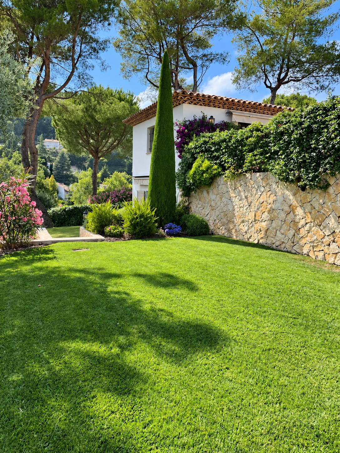 Une pelouse verte et soignée borde un mur en pierre couvert de végétation, avec un grand cyprès, des massifs fleuris et une villa blanche à toit de tuiles entourée de pins.