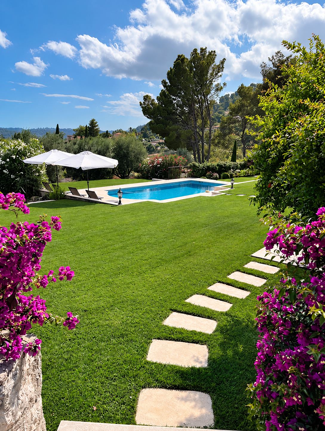 Un jardin soigné avec une pelouse verte traversée de dalles en pierre, une piscine rectangulaire, des parasols blancs et des bougainvilliers violets au premier plan.