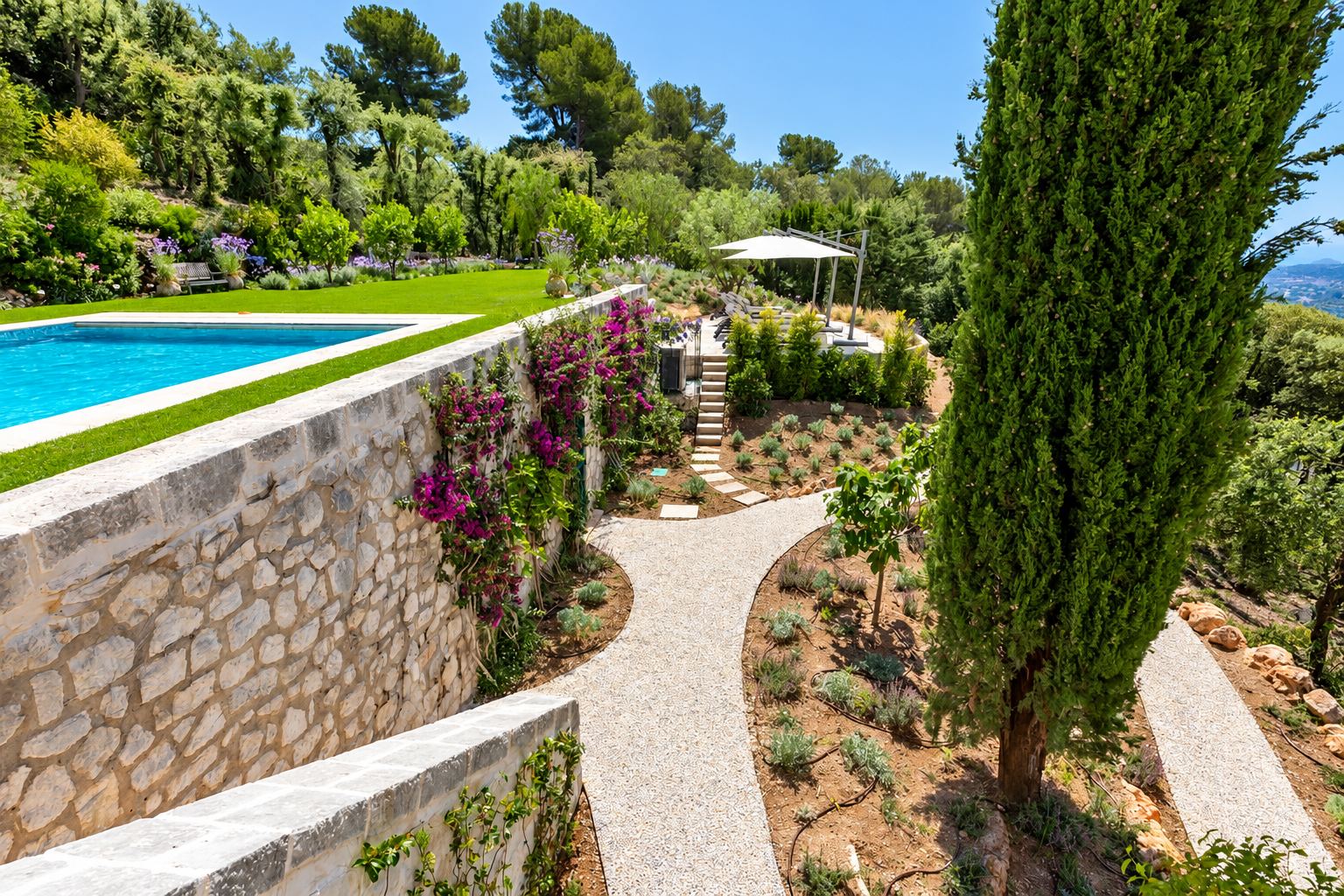 Jardin en terrasses avec piscine, haut mur de pierres sèches couvert de bougainvilliers, allée de gravier et cyprès sur une colline.
