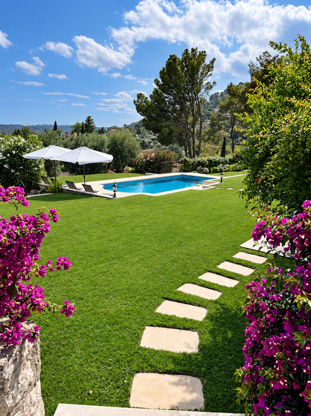 Un jardin méditerranéen avec une pelouse bien entretenue, un chemin de dalles en pierre, une piscine rectangulaire et des parasols blancs entourés de bougainvilliers violets.