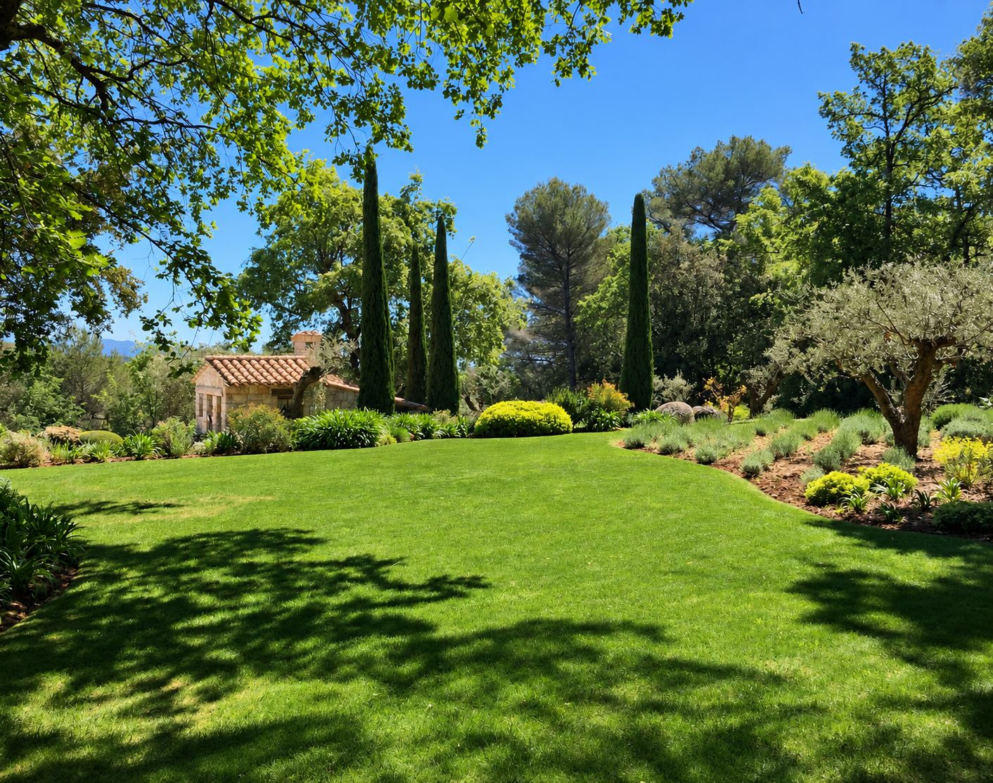 Un jardin méditerranéen soigné avec une grande pelouse verte, des cyprès, un olivier, des massifs de plantes et une petite maison en pierre au toit de tuiles.