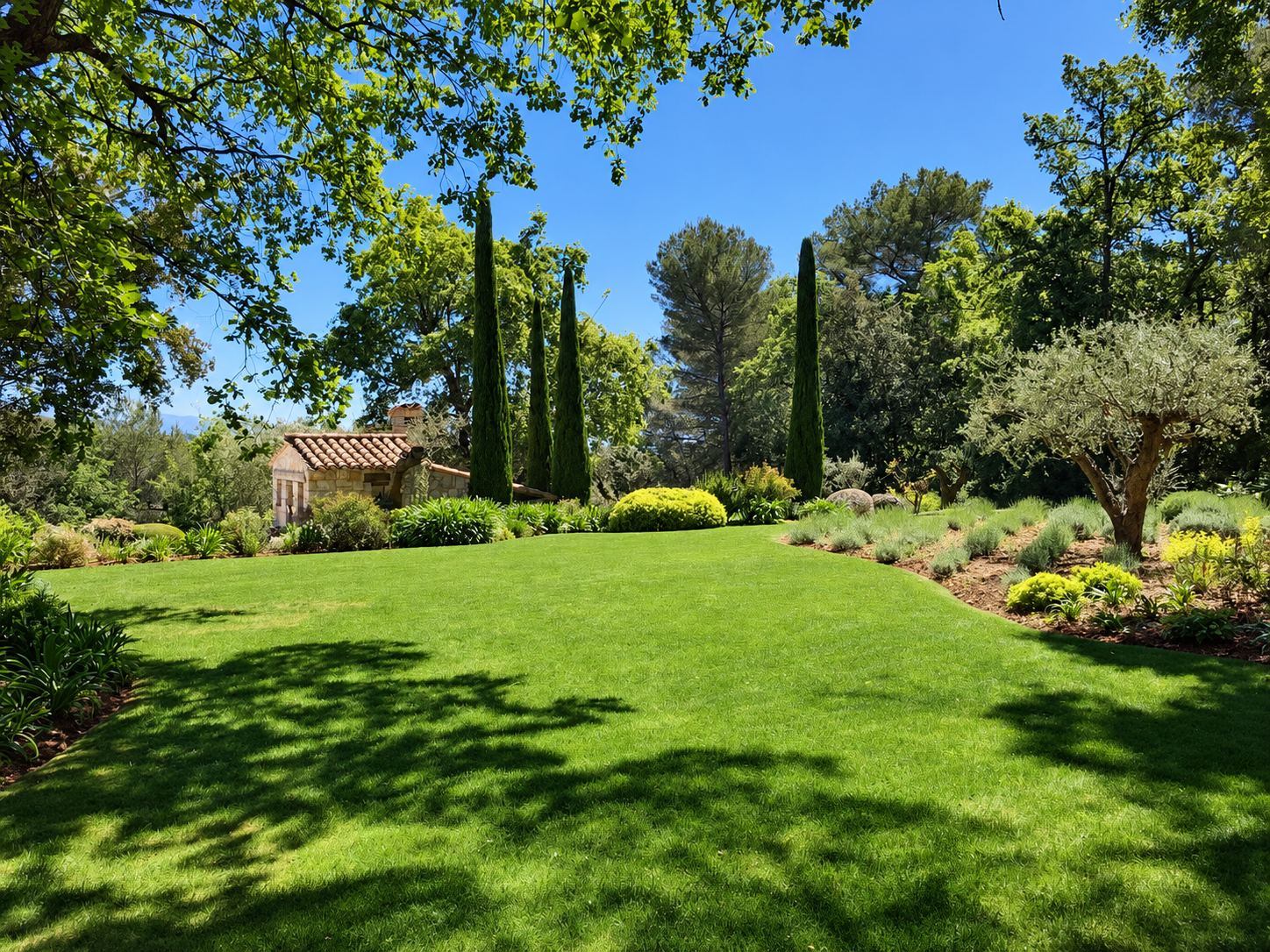 Un jardin méditerranéen soigné avec une grande pelouse verte, des cyprès, un olivier, des massifs de plantes et une petite maison en pierre sous un ciel bleu.