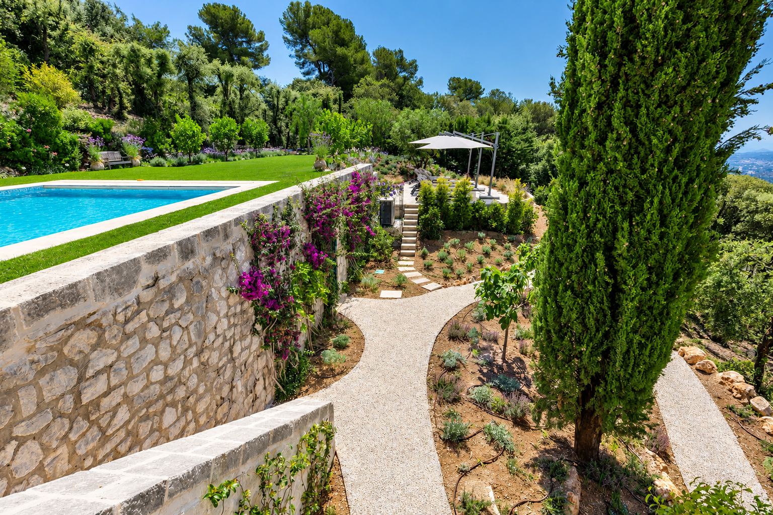 Jardin méditerranéen en terrasses avec une piscine bordée d'un mur en pierre, des allées de gravier, des massifs plantés et un grand cyprès.