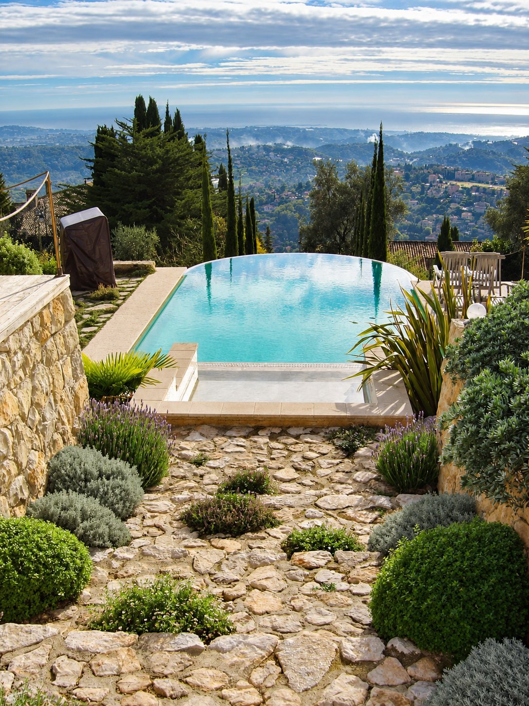Une piscine à débordement au bord arrondi surplombe un paysage de collines, avec un jardin méditerranéen et un sol en pierres plates au premier plan.