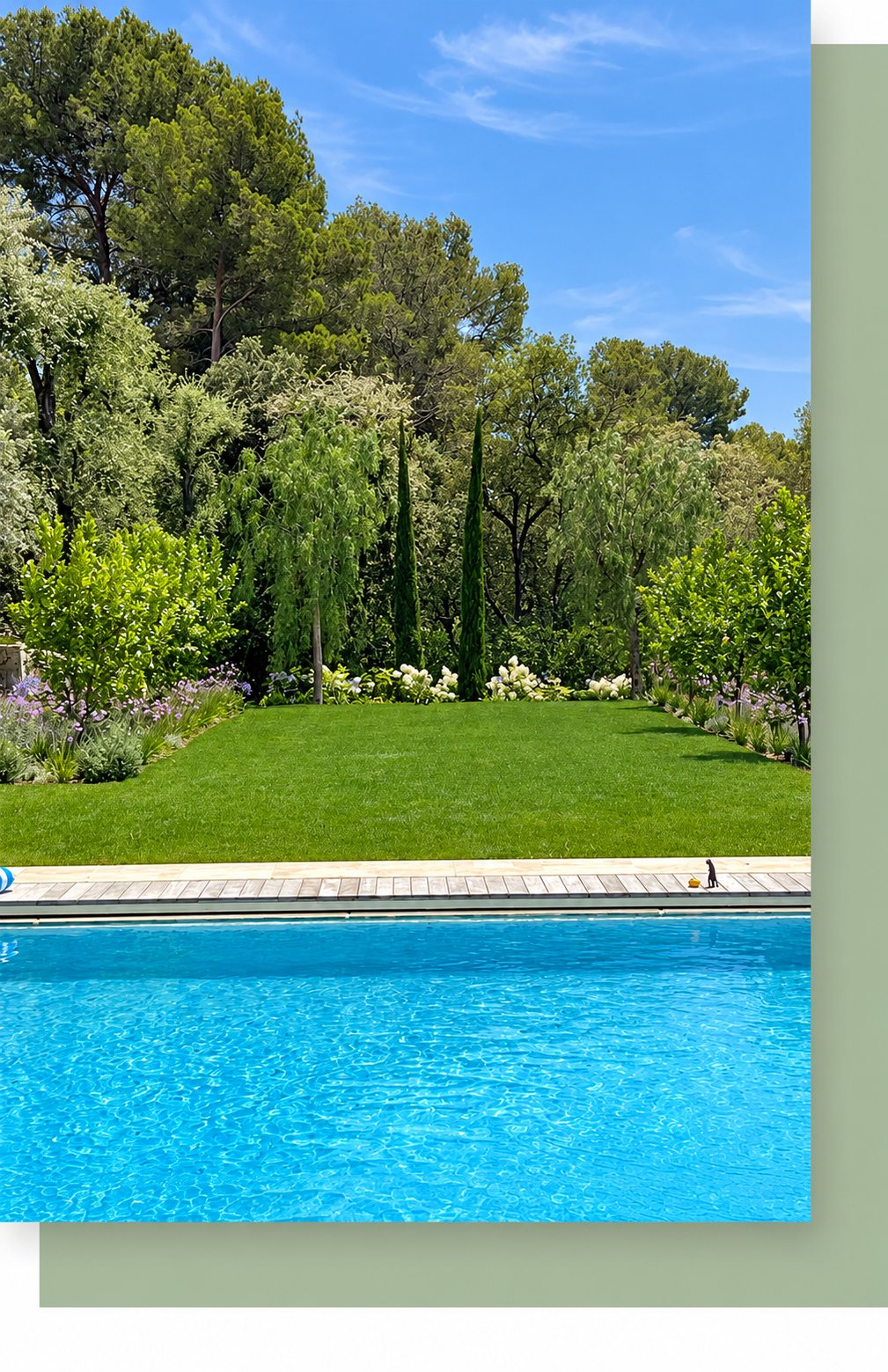 Une piscine d'eau turquoise borde une pelouse bien entretenue entourée de massifs fleuris, de cyprès et de grands arbres sous un ciel bleu.