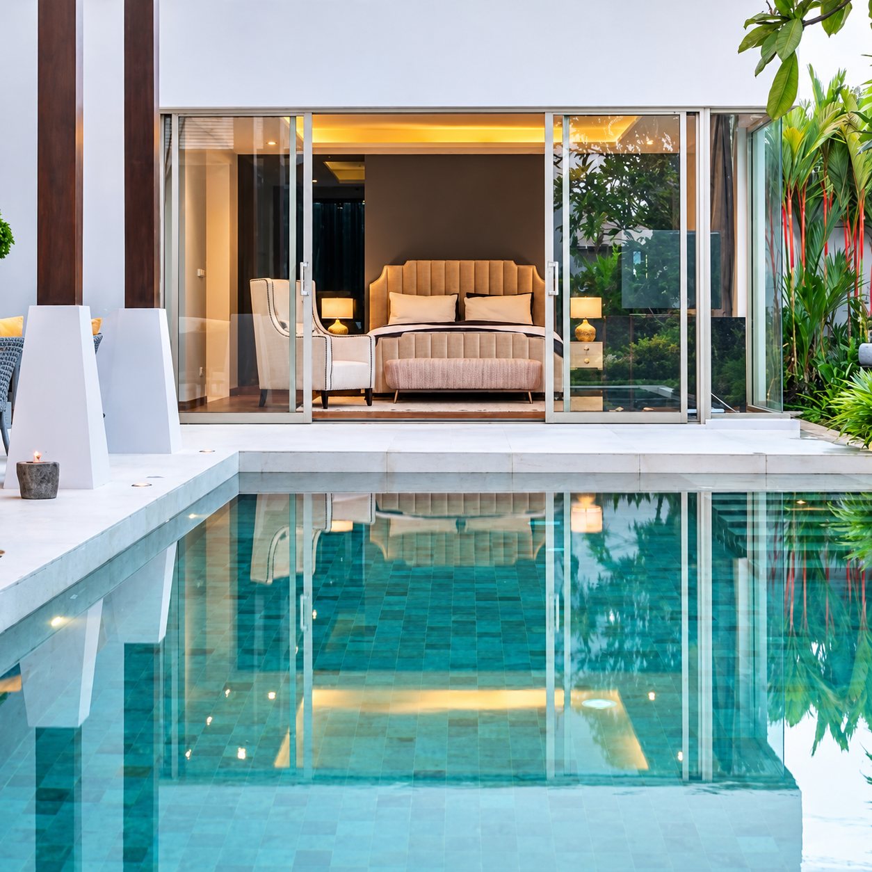 Une piscine au carrelage turquoise reflète les baies vitrées d'une chambre moderne avec un lit beige, entourée de plantes tropicales.