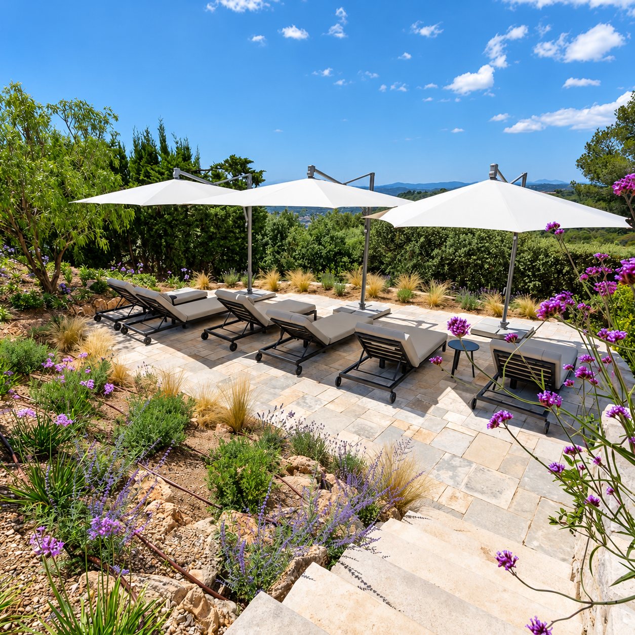 Une terrasse en pierre naturelle entourée d'un jardin méditerranéen fleuri, avec cinq bains de soleil gris et trois grands parasols blancs déportés sous un ciel bleu.