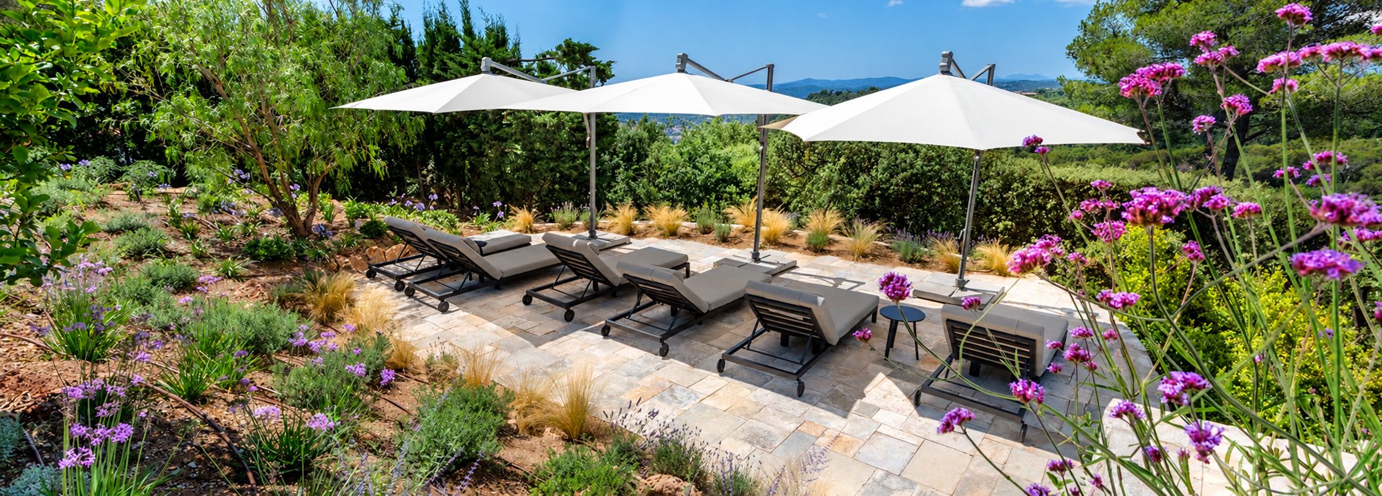 Une terrasse dallée de pierre entourée d'un jardin méditerranéen fleuri, équipée de bains de soleil gris et de grands parasols blancs déportés.